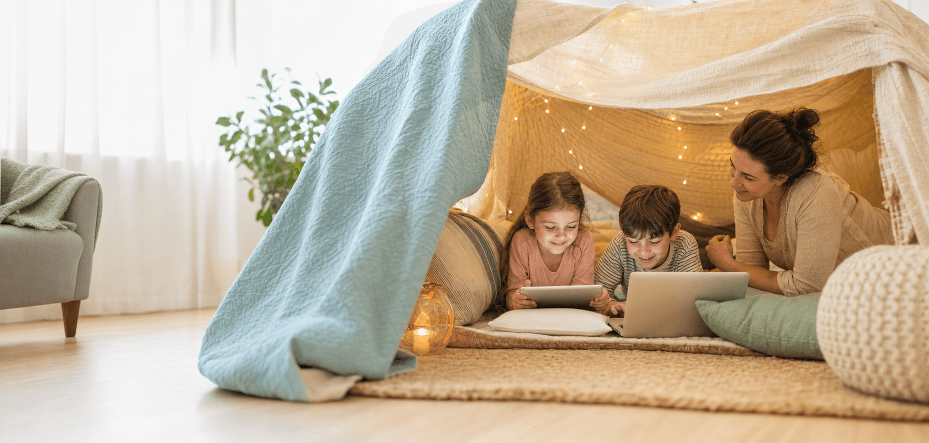 Children using devices inside a bright blanket fort while a parent looks on nearby.